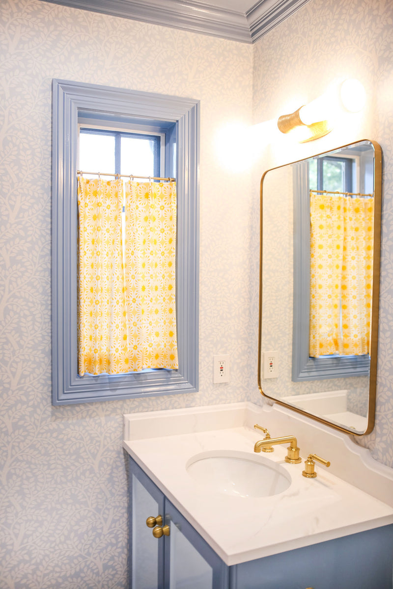 Bathroom with a two-tone blue vanity and brass accents, small window with a yellow floral cafe curtain